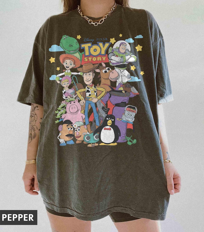 May include: Dark gray t-shirt featuring a colorful graphic of characters from the Disney Pixar movie, Toy Story. The design includes Woody, Buzz Lightyear, and other beloved characters. The shirt has a crew neck and short sleeves.