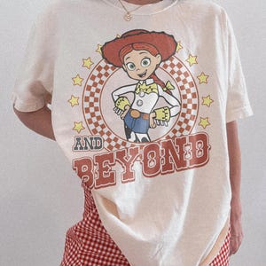 May include: An off-white short-sleeve t-shirt featuring a graphic of the character Jessie from Toy Story inside a checkered circle surrounded by yellow stars. The text AND BEYOND is printed in large, bold, reddish-brown letters below the graphic.