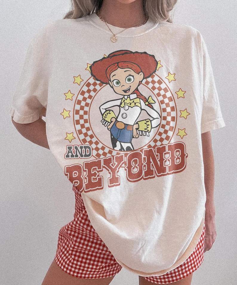 May include: An off-white short-sleeve t-shirt featuring a graphic of the character Jessie from Toy Story inside a checkered circle surrounded by yellow stars. The text AND BEYOND is printed in large, bold, reddish-brown letters below the graphic.