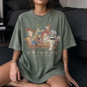 May include: An olive green short-sleeve t-shirt featuring a graphic print of Toy Story characters sitting on a couch. The text below the characters reads "F R I E N D S" and "SINCE 1995" in a light tan font. The shirt has a vintage-style wash.