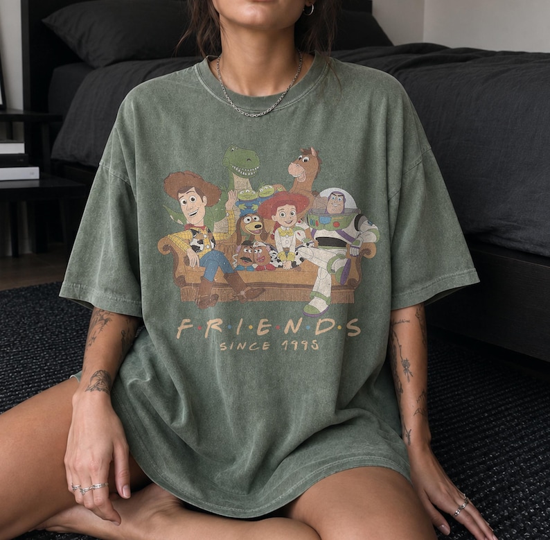 May include: An olive green short-sleeve t-shirt featuring a graphic print of Toy Story characters sitting on a couch. The text below the characters reads "F R I E N D S" and "SINCE 1995" in a light tan font. The shirt has a vintage-style wash.