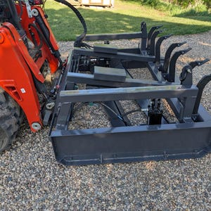 May include: An orange and black tractor with a gray grapple attachment. The grapple has curved tines and a lower bucket. The tractor is parked on a gravel surface.
