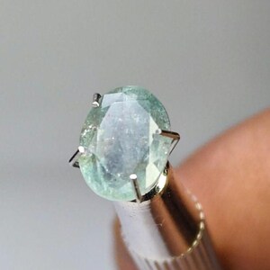 Puede incluir: Una gema ovalada de color verde claro, sujeta por un engaste de plata con garras. La gema tiene un aspecto ligeramente turbio y está sujeta por una herramienta metálica, probablemente para inspección o exhibición. El fondo es gris suave.