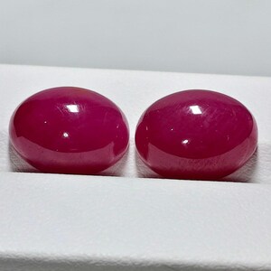 Op de afbeelding: Twee ovaalvormige, gepolijste edelstenen in een levendige, dieprode tint. De edelstenen hebben een glad, glanzend oppervlak dat licht reflecteert. De stenen zijn geplaatst tegen een witte achtergrond, waardoor hun kleur en vorm worden benadrukt. Dit kunnen robijnen of vergelijkbare edelstenen zijn.