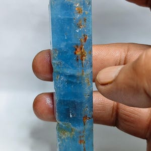May include: A long, translucent, light blue crystal specimen with a hexagonal prismatic shape. The surface features visible internal fractures and small, scattered patches of reddish-brown mineral inclusions along its vertical length.