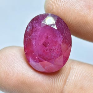 May include: An oval-shaped, faceted ruby gemstone with a deep pinkish-red hue. The stone features a polished surface with visible internal inclusions and a distinct light reflection on its top facet, showcasing a translucent and vibrant appearance.