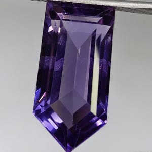 May include: A faceted, elongated purple gemstone held by silver tweezers. The gem has a rectangular shape with angled edges and a pointed bottom. The facets reflect light, creating a sparkling effect. The background is a neutral gray.