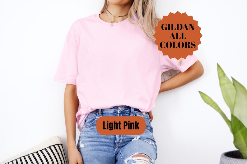 May include: A light pink t-shirt with the text "Light Pink" on the front. The shirt is a solid color and is being worn with blue jeans. A brown and orange sticker on the shirt says "GILDAN ALL COLORS".