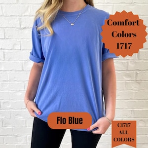 May include: A blue Comfort Colors t-shirt with rolled-up sleeves. The image displays the text "Flo Blue", "Comfort Colors 1717", and "C1717 ALL COLORS". The shirt is worn by a person in front of a white brick wall.