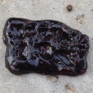 May include: A close-up of a dark, shiny, irregularly shaped object with a textured surface. The object appears to be made of a viscous substance, possibly a type of resin or tar.