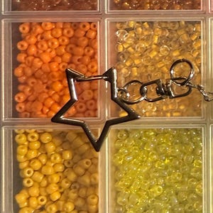 Puede incluir: Un organizador de pl&aacute;stico transparente lleno de peque&ntilde;as cuentas de colores en tonos naranja, amarillo y dorado. Un dije plateado en forma de estrella con una cadena est&aacute; unido al organizador, lo que sugiere un proyecto de joyer&iacute;a o manualidades.