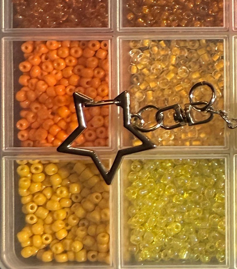 Puede incluir: Un organizador de pl&aacute;stico transparente lleno de peque&ntilde;as cuentas de colores en tonos naranja, amarillo y dorado. Un dije plateado en forma de estrella con una cadena est&aacute; unido al organizador, lo que sugiere un proyecto de joyer&iacute;a o manualidades.