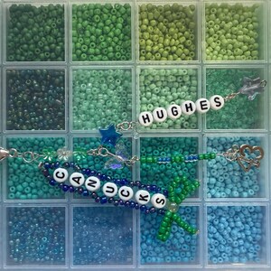 Puede incluir: Un organizador de pl&aacute;stico transparente lleno de cuentas de varios tonos de verde y azul. Se ven dos llaveros, uno con la palabra "CANUCKS" y otro con "HUGHES", ambos con adornos decorativos.