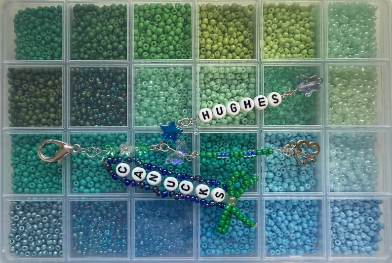 Puede incluir: Un organizador de pl&aacute;stico transparente lleno de cuentas de varios tonos de verde y azul. Se ven dos llaveros, uno con la palabra "CANUCKS" y otro con "HUGHES", ambos con adornos decorativos.