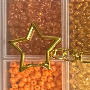 Puede incluir: Un dije dorado en forma de estrella con un cierre de mosquet&oacute;n, sobre un fondo de peque&ntilde;as cuentas de colores en un recipiente dividido. Las cuentas son en tonos naranja, dorado y marr&oacute;n.