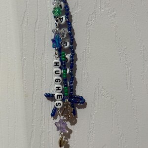 Puede incluir: Un llavero de cuentas hecho a mano con el nombre "HUGHES" escrito con cuentas de letras blancas. El llavero presenta cuentas azules, verdes y transparentes, junto con dijes de estrella y coraz&oacute;n. Las cuentas est&aacute;n ensartadas en una cadena plateada.