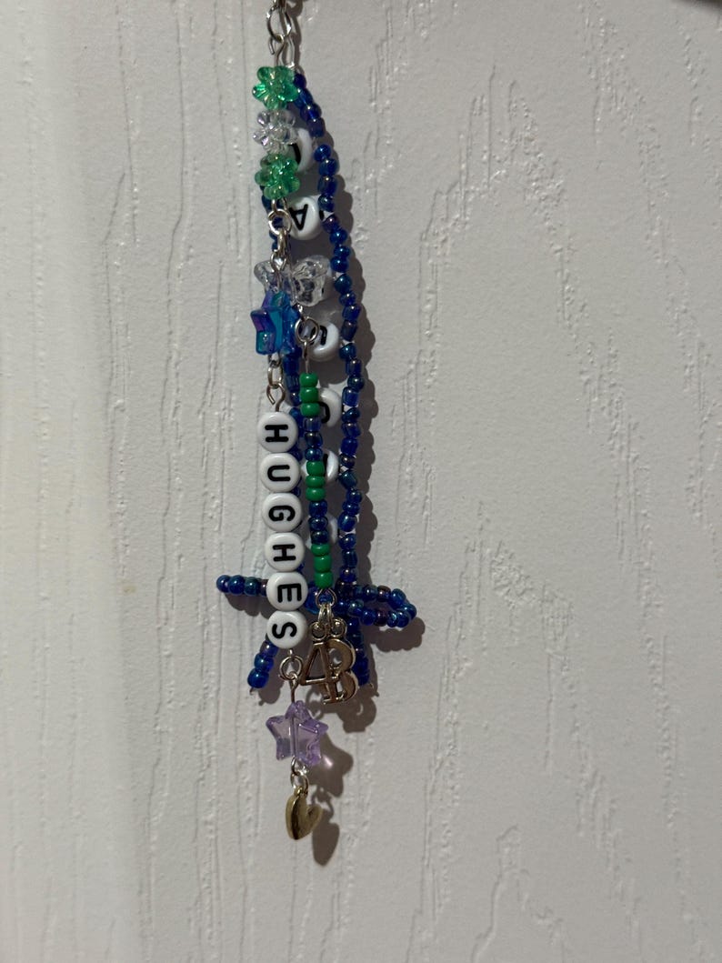 Puede incluir: Un llavero de cuentas hecho a mano con el nombre "HUGHES" escrito con cuentas de letras blancas. El llavero presenta cuentas azules, verdes y transparentes, junto con dijes de estrella y coraz&oacute;n. Las cuentas est&aacute;n ensartadas en una cadena plateada.