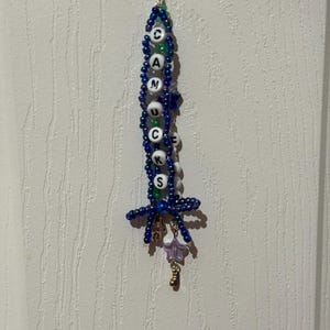 Puede incluir: Un llavero de cuentas con la palabra "CANUCKS" escrita con cuentas de letras blancas. El llavero presenta cuentas azules y verdes, un peque&ntilde;o dije de estrella y un dije dorado. El llavero cuelga contra una pared blanca texturizada.