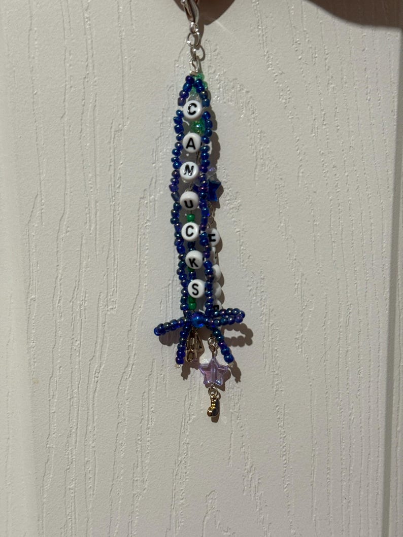 Puede incluir: Un llavero de cuentas con la palabra "CANUCKS" escrita con cuentas de letras blancas. El llavero presenta cuentas azules y verdes, un peque&ntilde;o dije de estrella y un dije dorado. El llavero cuelga contra una pared blanca texturizada.