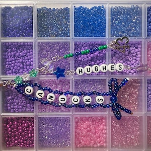 Puede incluir: Un organizador de pl&aacute;stico transparente lleno de cuentas de varios tonos de morado, azul y rosa. Se ven dos pulseras de cuentas, una con la palabra "CANUCKS" y la otra con "HUGHES". Las pulseras tienen dijes y un lazo.