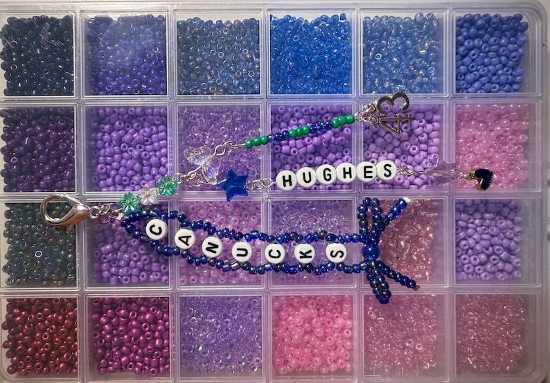 Puede incluir: Un organizador de pl&aacute;stico transparente lleno de cuentas de varios tonos de morado, azul y rosa. Se ven dos pulseras de cuentas, una con la palabra "CANUCKS" y la otra con "HUGHES". Las pulseras tienen dijes y un lazo.
