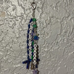 Puede incluir: Un llavero de cuentas hecho a mano con las palabras "CANUCKS" y "HUGHES" deletreadas en cuentas de letras blancas. El llavero presenta cuentas azules, verdes y moradas, junto con dijes de estrellas y mariposas, y un cierre plateado.
