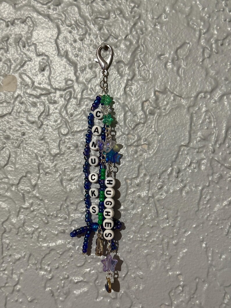 Puede incluir: Un llavero de cuentas hecho a mano con las palabras "CANUCKS" y "HUGHES" deletreadas en cuentas de letras blancas. El llavero presenta cuentas azules, verdes y moradas, junto con dijes de estrellas y mariposas, y un cierre plateado.