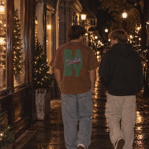 May include: A brown short-sleeved t-shirt featuring the green block-lettered text BABY with the word Santa written in pink cursive script across the center. The shirt is worn with loose-fitting blue denim jeans and white sneakers.