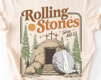 May include: Cream-colored t-shirt featuring a graphic with the text "Rolling Stones" in brown and orange. The graphic depicts a tomb with a large stone, crosses, and pine trees. The text "Since AD 33" is also included.
