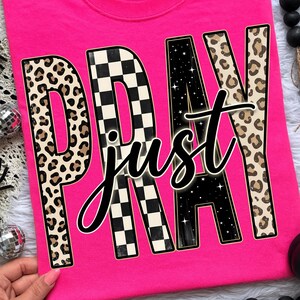 Könnte beinhalten: Ein leuchtend pinkfarbenes T-Shirt mit dem Schriftzug "PRAY just" in einem auffälligen Grafikdesign. Die Buchstaben sind mit Leopardenmuster, einem schwarz-weißen Schachbrettmuster und einem Sternennacht-Design verziert.