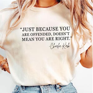 May include: Cream-colored t-shirt featuring the quote "JUST BECAUSE YOU ARE OFFENDED, DOESN'T MEAN YOU ARE RIGHT." in black text, with "-Charlie Kirk" below. The shirt has short sleeves.