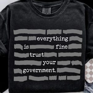 Puede incluir: Sudadera negra de cuello redondo con el texto blanco "everything is fine trust your government". El texto está dispuesto horizontalmente con un estilo vintage y desgastado. La sudadera es de color oscuro y descolorido.