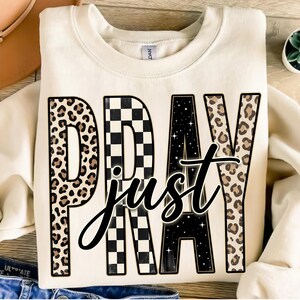 Może przedstawiać: Kremowa bluza z napisem "PRAY just PRAY" w odważnym designie. Litery mają różne wzory: panterkę, czarno-białą szachownicę i gwiaździste niebo. Codzienny element garderoby o tematyce religijnej.