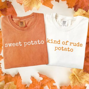 May include: Two folded t-shirts, one orange and one white, with text. The orange shirt says "sweet potato" in white letters. The white shirt says "kind of rude potato" in orange letters. Autumn leaves surround the shirts.