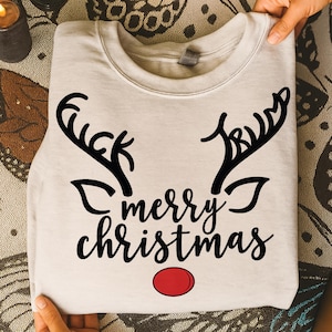 以下が含まれることがあります： 黒いトナカイの角のデザインと「merry christmas」の文字が書かれた、折りたたまれた薄いベージュのスウェットシャツ。赤い円はトナカイの鼻を表しています。