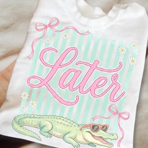 Peut inclure: T-shirt blanc avec un alligator de dessin animé portant des lunettes de soleil roses. Le mot "Later" est écrit en rose sur un fond à rayures vertes menthe et blanches, avec des marguerites et des nœuds roses.