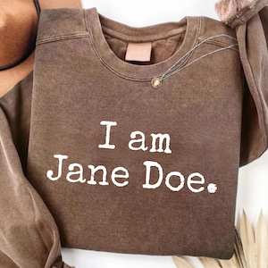 Puede incluir: Una sudadera marrón de cuello redondo con el texto blanco "I am Jane Doe." estampado en la parte delantera. La prenda tiene mangas largas con cuello, puños y dobladillo acanalados, y presenta una textura de tela desgastada y vintage.