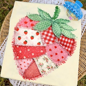 May include: A handmade strawberry-shaped patchwork design with a cream-colored background. The strawberry is composed of various red and pink fabrics, including gingham, floral, and heart patterns. Green leaves and a blue hair clip complete the image.