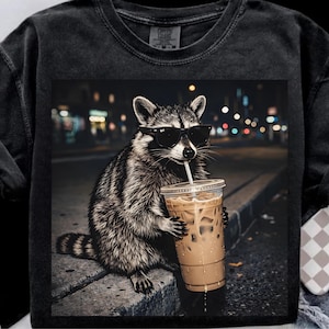 May include: Black Comfort Colors sweatshirt featuring a raccoon wearing sunglasses and drinking iced coffee. The raccoon is sitting on a curb with a city street background. The image is a graphic design.