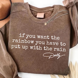 Könnte beinhalten: Ein braunes Sweatshirt mit Rundhalsausschnitt und weißem Text "if you want the rainbow you have to put up with the rain", gefolgt von der Signatur "Dolly" in Schreibschrift. Das Kleidungsstück hat ein verwaschenes Vintage-Finish.