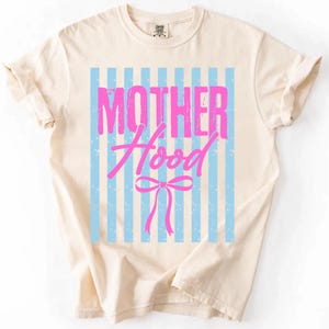 May include: Cream-colored t-shirt with the word "MOTHER" above "Hood" in pink, set against a light blue striped background. A pink bow is below "Hood". The shirt has short sleeves and a crew neck.