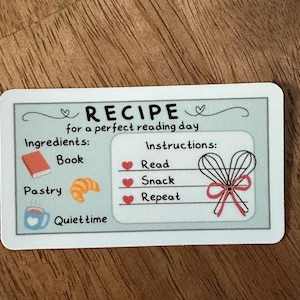 May include: A rectangular sticker with a light blue background features a recipe for a perfect reading day. The recipe includes ingredients like a book, pastry, and quiet time, with instructions to read, snack, and repeat.