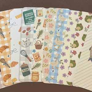 May include: A collection of decorative bookmarks with various patterns and illustrations. Designs include gingham, croissants, jars of jam, ducks, frogs, and recipe-themed elements. One bookmark reads "gone reading..."