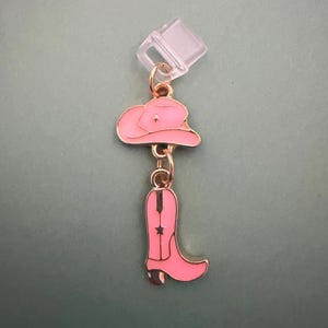 May include: A pink cowboy hat and boot charm with gold-coloured accents. The charm is attached to a clear plastic clip. The hat has a star detail, and the boot has a black stripe and a star. The charm is likely for a keychain or accessory.