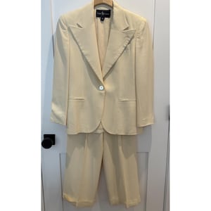 May include: A cream-colored Ralph Lauren suit, including a blazer and matching pants. The blazer has a single button closure and notched lapels. The pants are a cropped style. The suit is hanging on a wooden hanger.