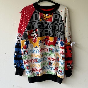 May include: A patchwork sweatshirt featuring Mickey Mouse and friends. The design incorporates various patterns, including polka dots, stars, and the words "Mickey" and "Key & Go." The sleeves and body are a mix of red, white, blue, and yellow panels. Black cuffs and neckline complete the look.