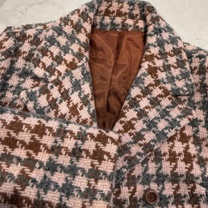 May include: A close-up of a houndstooth patterned coat in shades of pink, brown, and gray. The coat has a notched collar, a brown button, and a brown lining. The fabric has a woven texture.