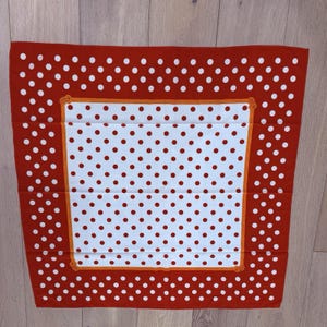 May include: A square scarf with a red border and white polka dots. The center features a white background with red polka dots, framed by an orange border. The scarf is folded and lies flat on a wooden surface.