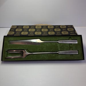 May include: A vintage carving set, including a serrated knife and a two-tine fork, presented in a black box with a green velvet lining. The box lid features a gold and black patterned design. The knife and fork have silver-toned metal blades and handles.