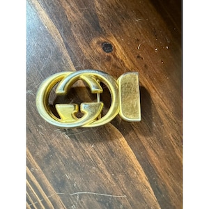 May include: A gold-colored belt buckle with a double-G logo. The buckle has a rectangular bar on one side for the belt attachment. The logo is a stylized interlocking design.